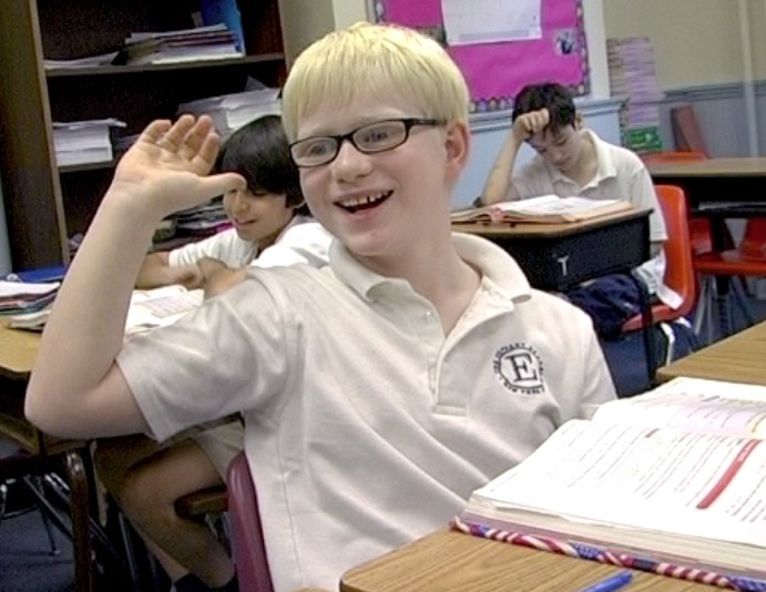 “Emmet in Going Blind” - a boy with blond hair and glasses sits at a small classroom desk.  The boy is smiling enthusiastically and raising his hand to be called on by the teacher who is not in the photo.
