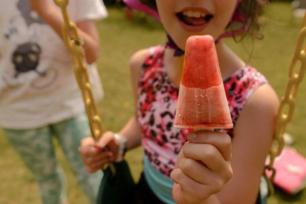 a smiling child is sitting on a swing and holding a popsicle out toward the camera 