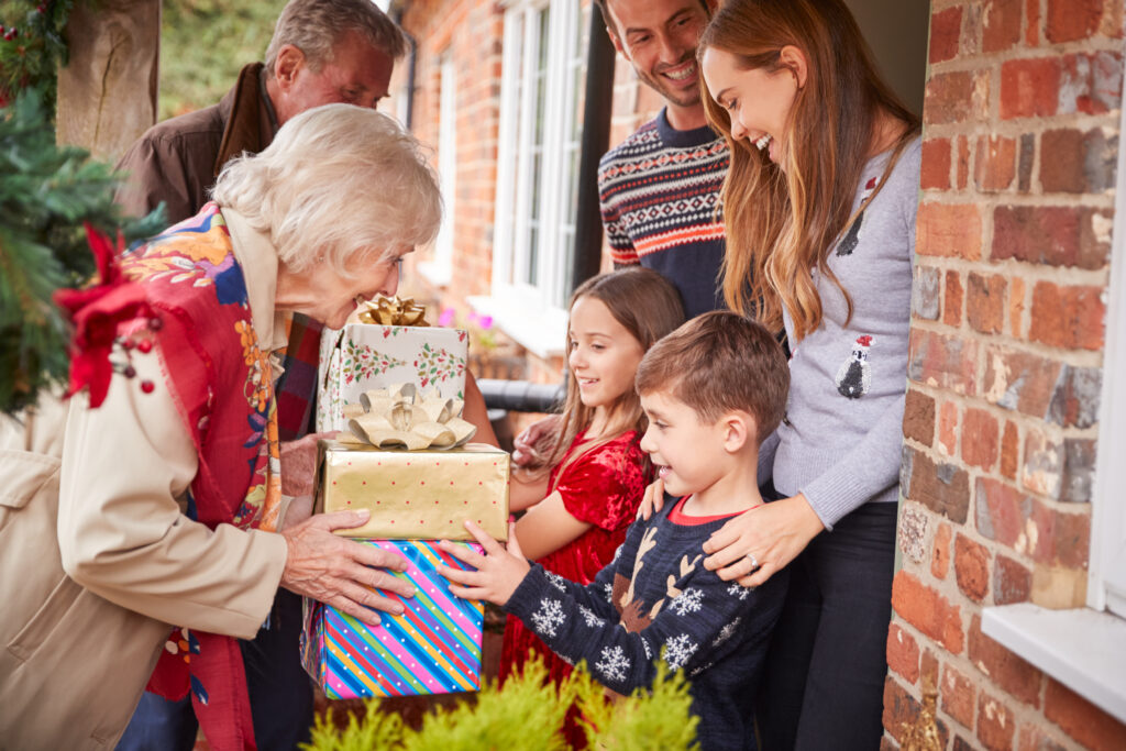 Grandparents are presenting gifts to a mother, father and their two children at the door to their home. 