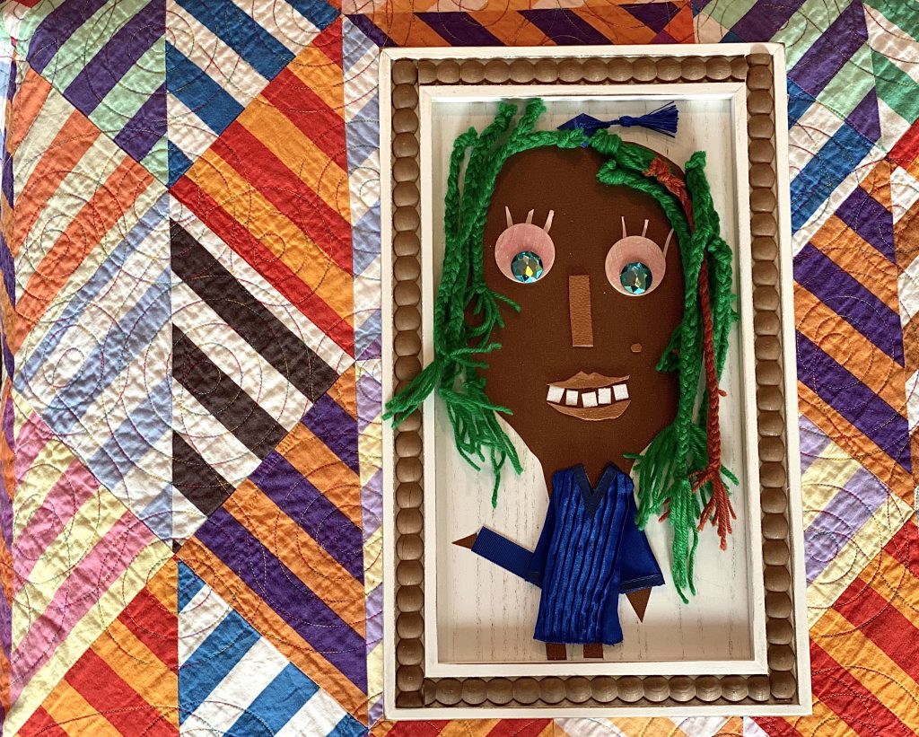 multi-textured, collage self-portrait with cardstock face, wooden eyes, leather lips, ribbon nose, and braided yarn hair  