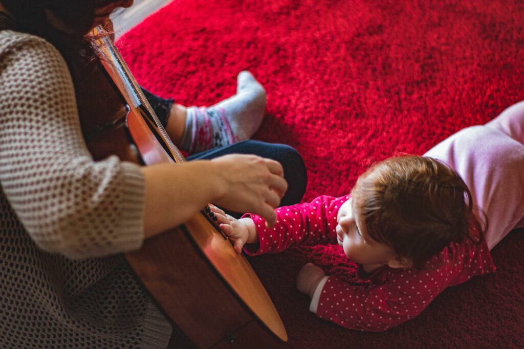 An adult sitting on the floor holding a guitar while a toddler holds his hands against the guitar's strings. 