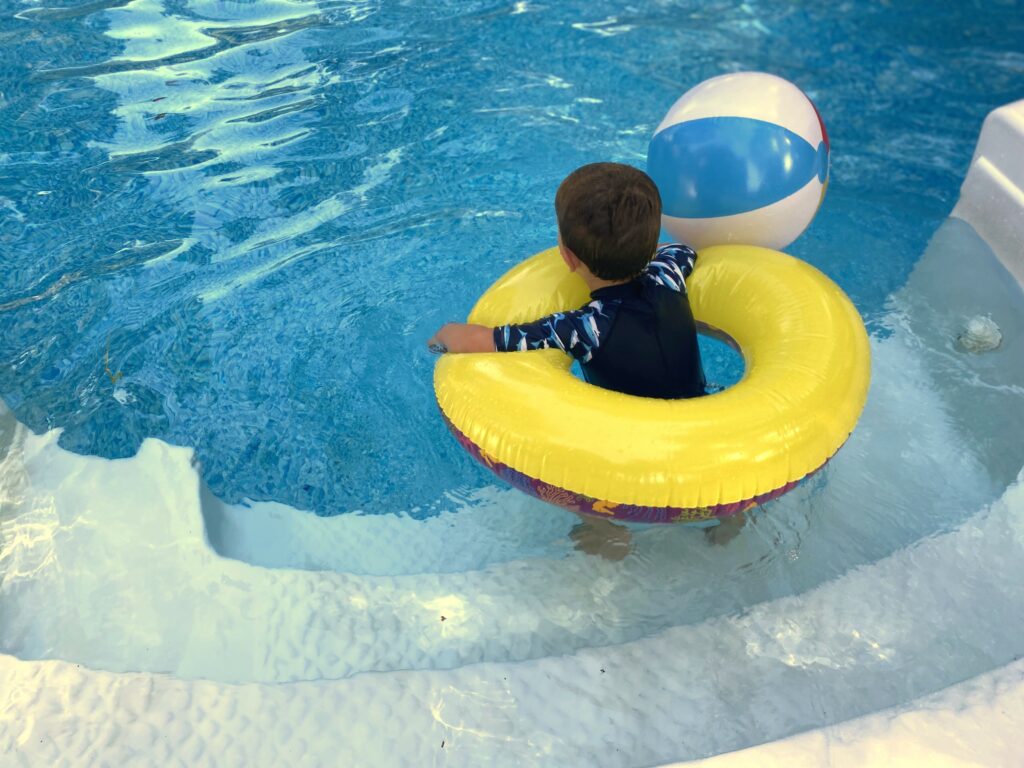 A preschool aged boy is standing on the steps of a pool with a yellow ring float around him and a striped beach ball next to him.