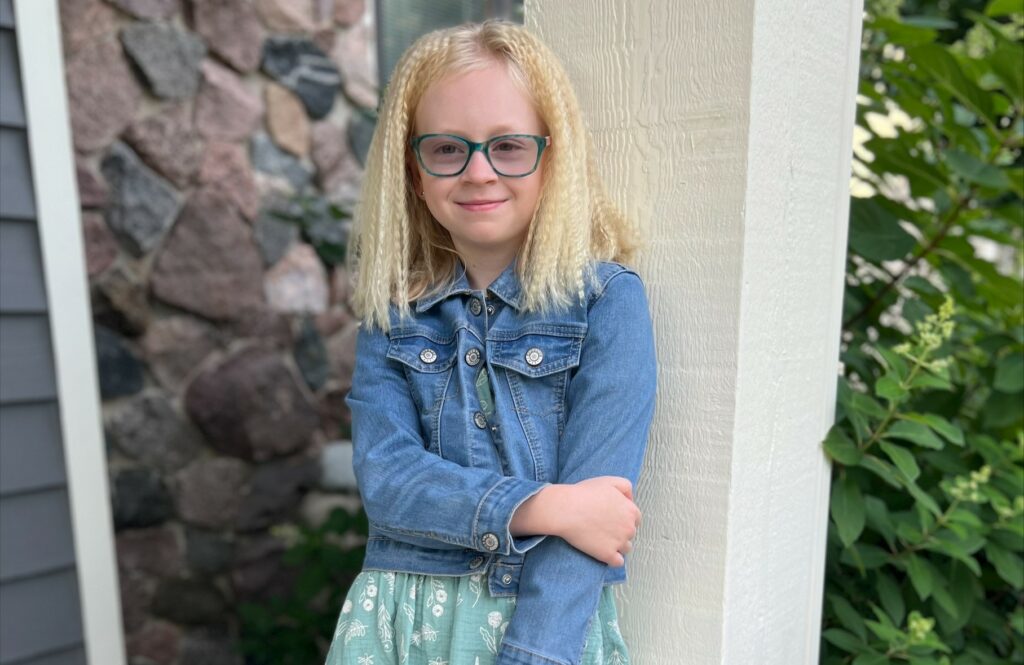 Ally, with light blond hair and teal eyeglasses, clutches her left arm with her right hand