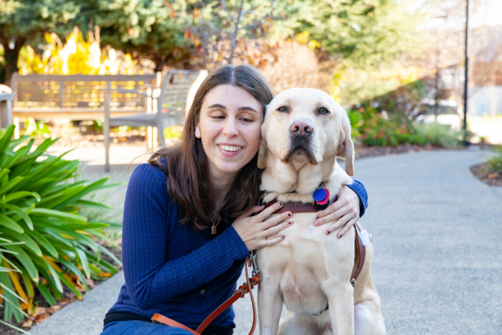 Young adult with her arm around a wheat-colored dog guide in harness