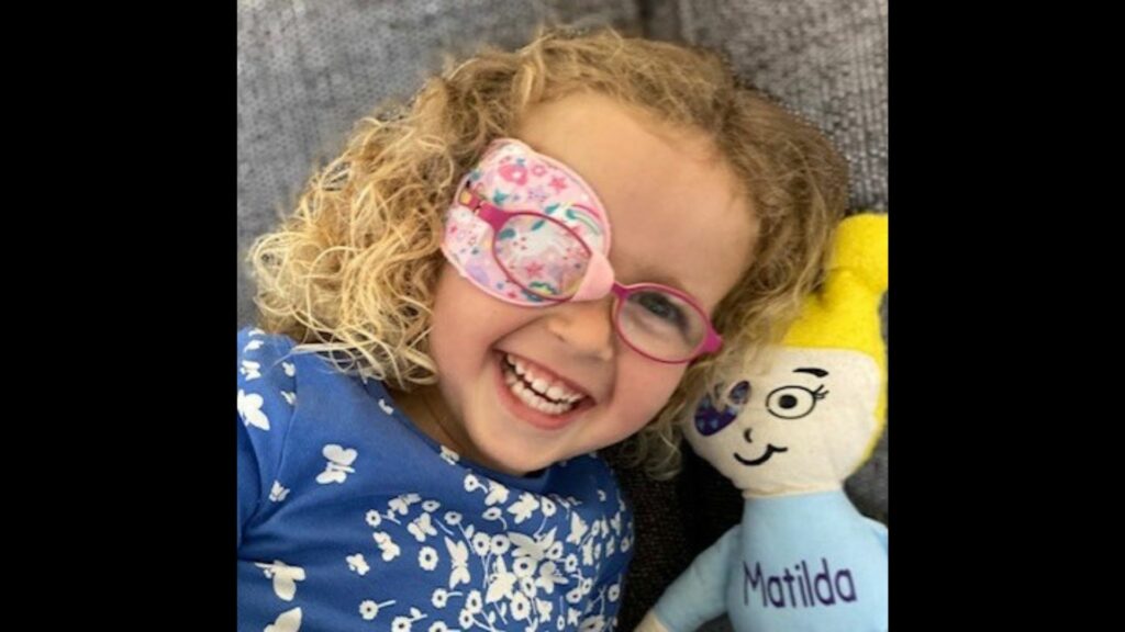 young girl wearing pink eyeglasses and a colorful eyepatch sitting next to a cloth doll with an eye patch and "Matilda" embroidered
