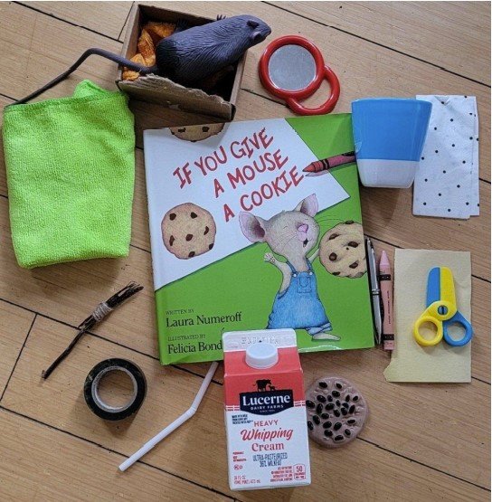 On the center of the picture, there is a book called “ If You Give A Mouse A Cookie”. There are some objects placed around the book. From the top right: Mouse toy, paper box (represents a bed), mirror, cup, napkin, pen, crayon, scissors, paper, toy cookie, whipping cream carton (represents milk), straw, tape, broom, cloth.