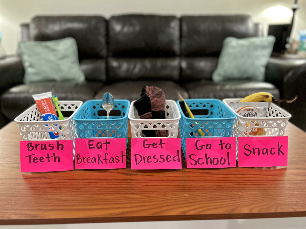 Photo shows five calendar boxes in alternating colors with bold print labels. Real items are included in each box. The labels read from left to right: Brush Teeth, Eat Breakfast, Get Dressed, Go to School, Snack.
