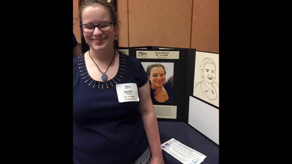 Jessica is standing and smiling toward the camera. Next to her is a poster board. On the left side, a picture of Jessica is visible along with a printed biography. On the right side of the poster, there is a tactile image of Jessica and a brailled biography underneath it.