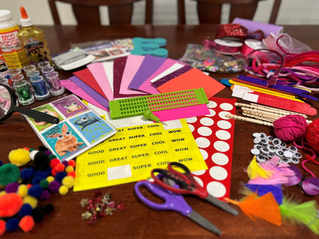 A variety of craft materials displayed on a table. Items include bold print and tactile stickers, a slate and stylus, scissors, store-bought valentine cards, feathers, yarn, google eyes, fuzzy pom-poms, craft sticks, pipe cleaners, ribbon, glitter, and glue. 