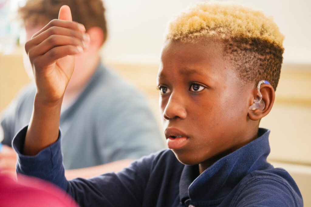 A boy wearing a hearing aid raising his hand in a classroom setting.