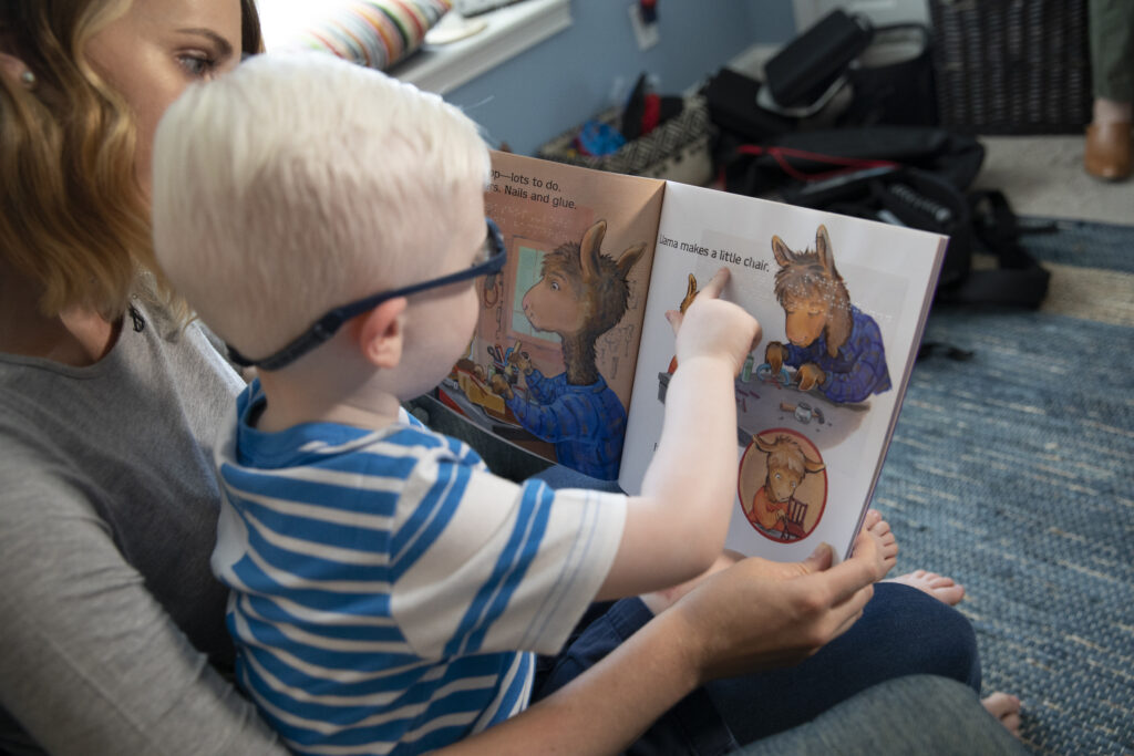 A young boy sitting in his mom's lap reading a book and pointing to the photo. 