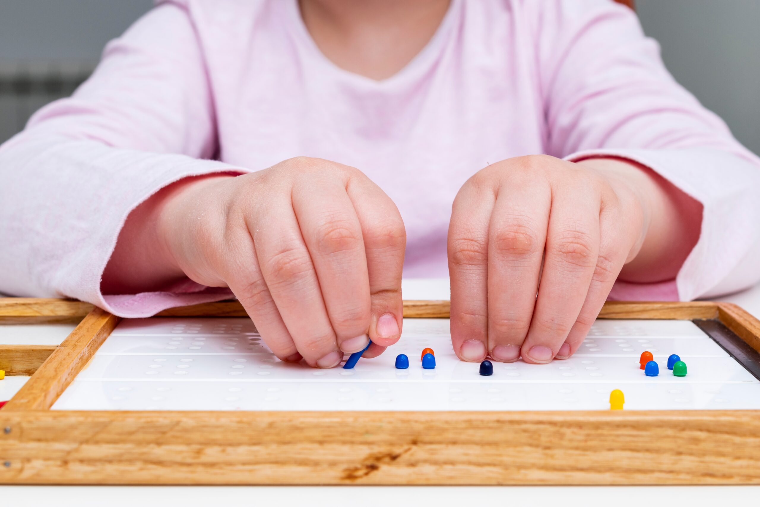 A child sitting at a table with her hands placing small pegs into braille cells. 