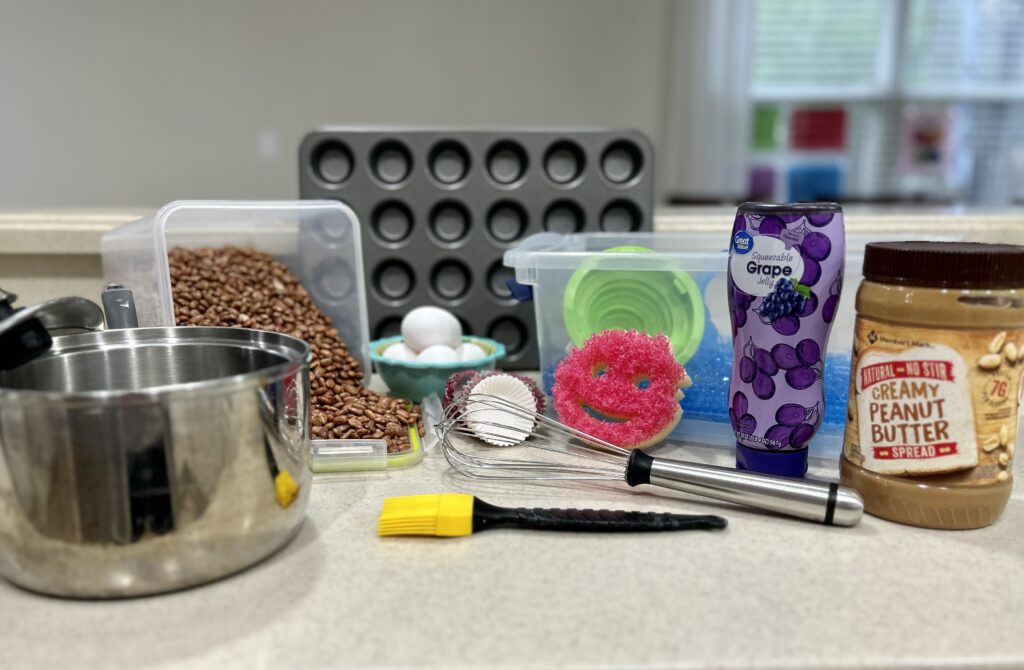 The photo shows various household items that can be used for sensory efficiency activities. Items include a container with dried beans, a bin of water beads, a pot with a lid, a whisk, a dish scrubber, a jar of peanut butter, a few eggs in a bowl, a jar of jelly, baking cups, and a muffin pan.