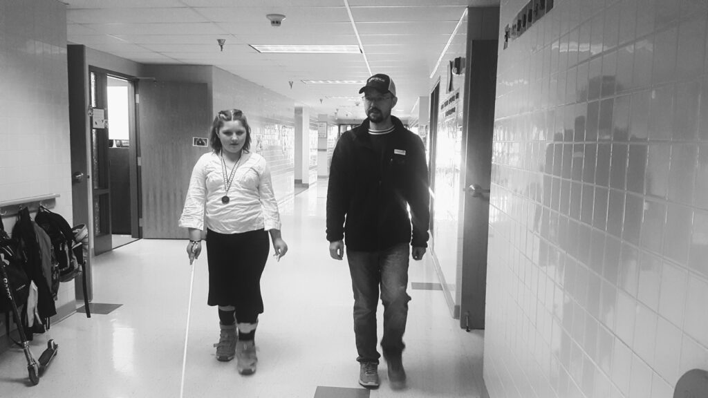A teen using a white cane and an adult walk down a hallway